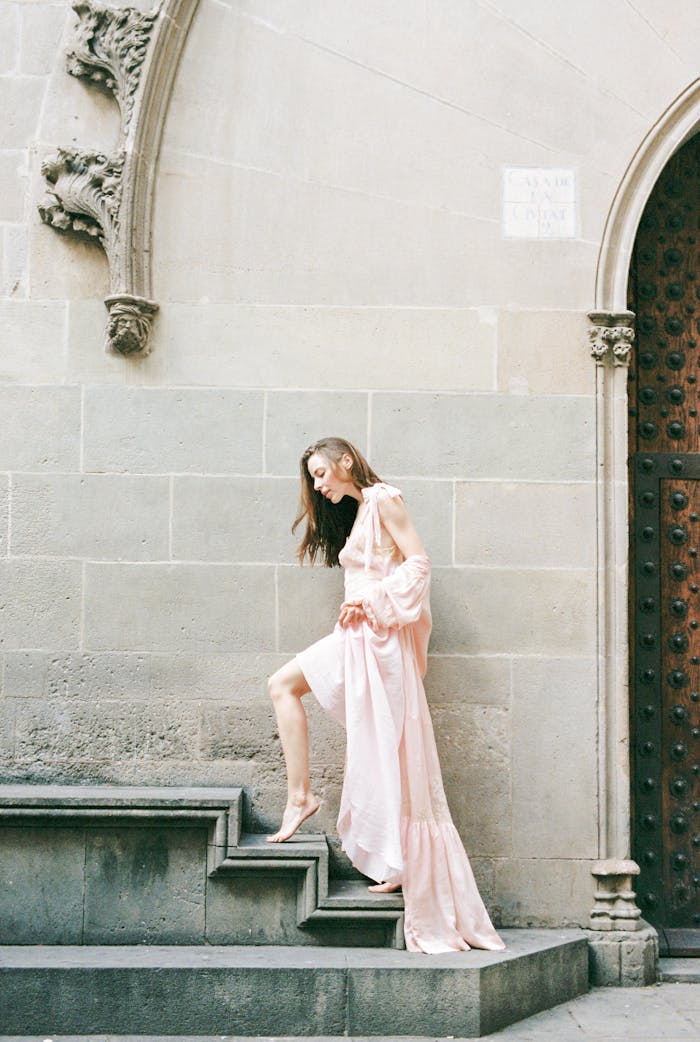 A graceful woman in a pink dress ascends stone steps beside an ornate wall.